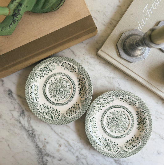 Two small decorative plates with a green and white floral toile pattern, arranged on a white marble surface next to books and a candlestick.
